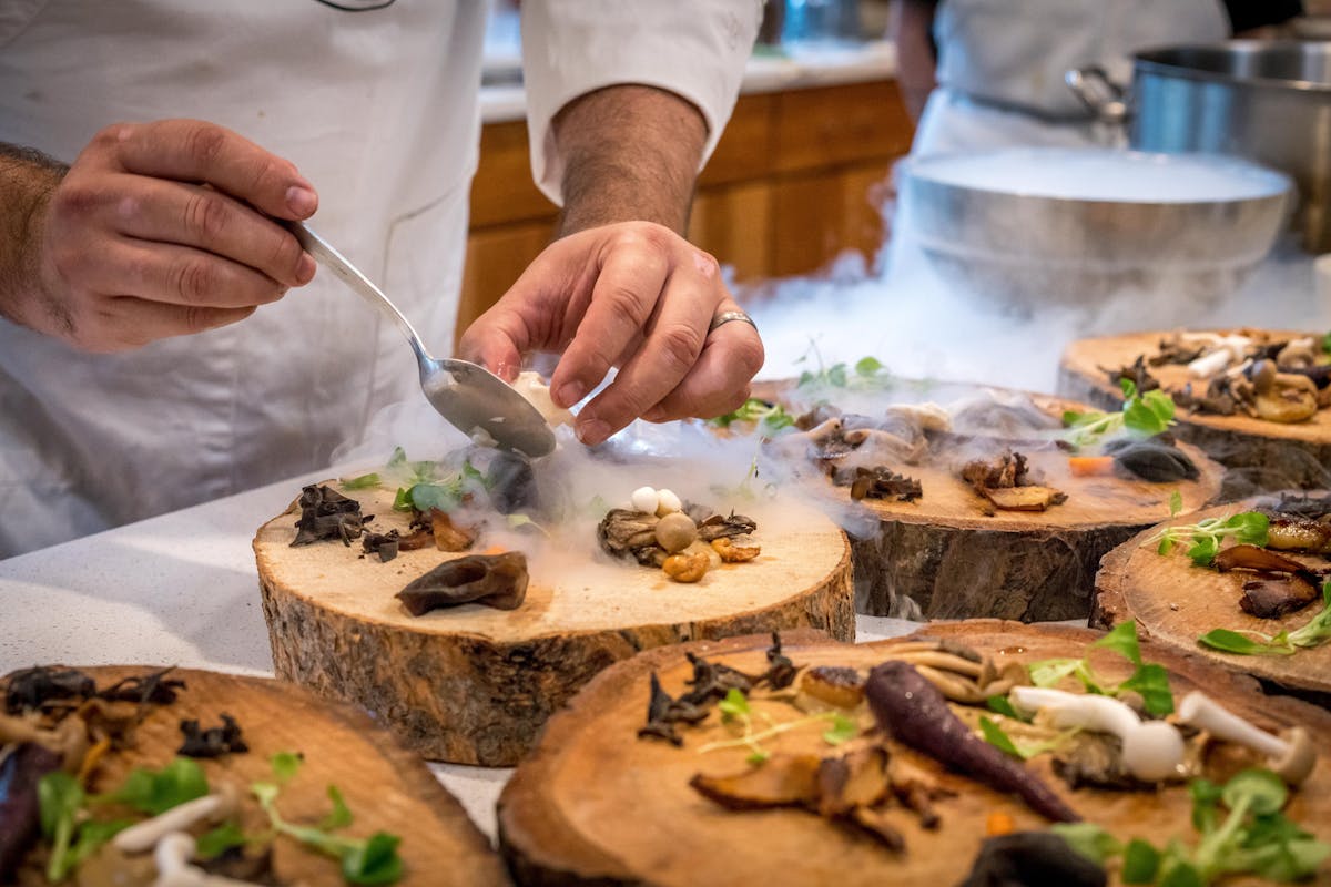 Chef in whites plates mushrooms onto log-slice boards as dry-ice mist rolls across the counter